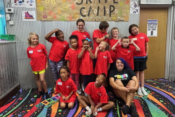Group of kids in red shirts and an adult posing playfully under a 'Skate Camp' sign indoors.