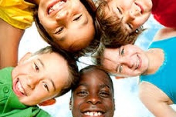 Five smiling children in colorful shirts lie in a circle, heads touching, against a blue sky background.