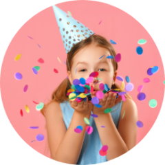 Child in party hat blowing colorful confetti on pink background.