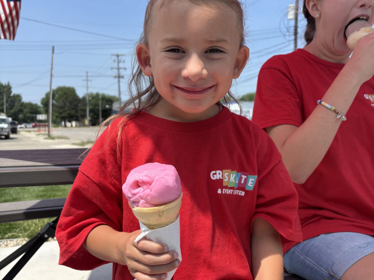 Child in red shirt holding ice cream under a pink umbrella with another person in background.