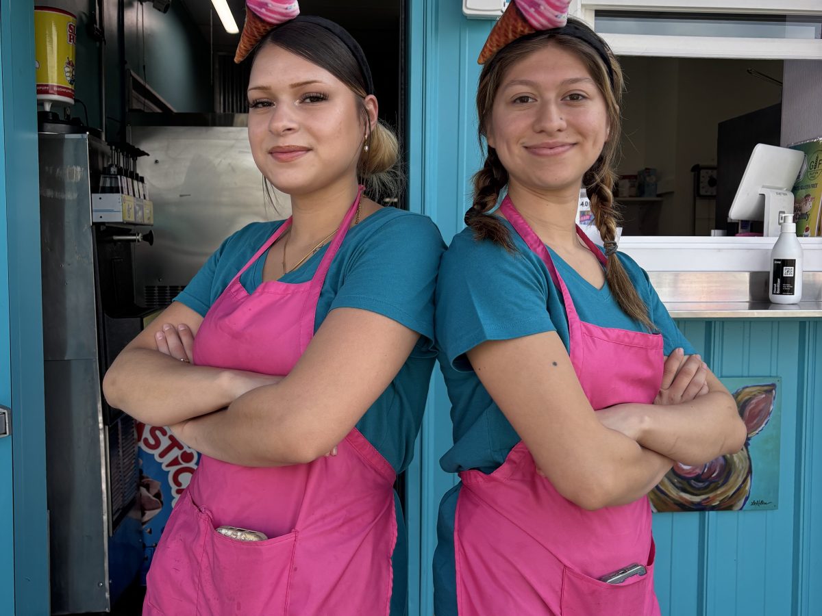 Two smiling workers in pink aprons with ice cream hats in front of a colorful shop.