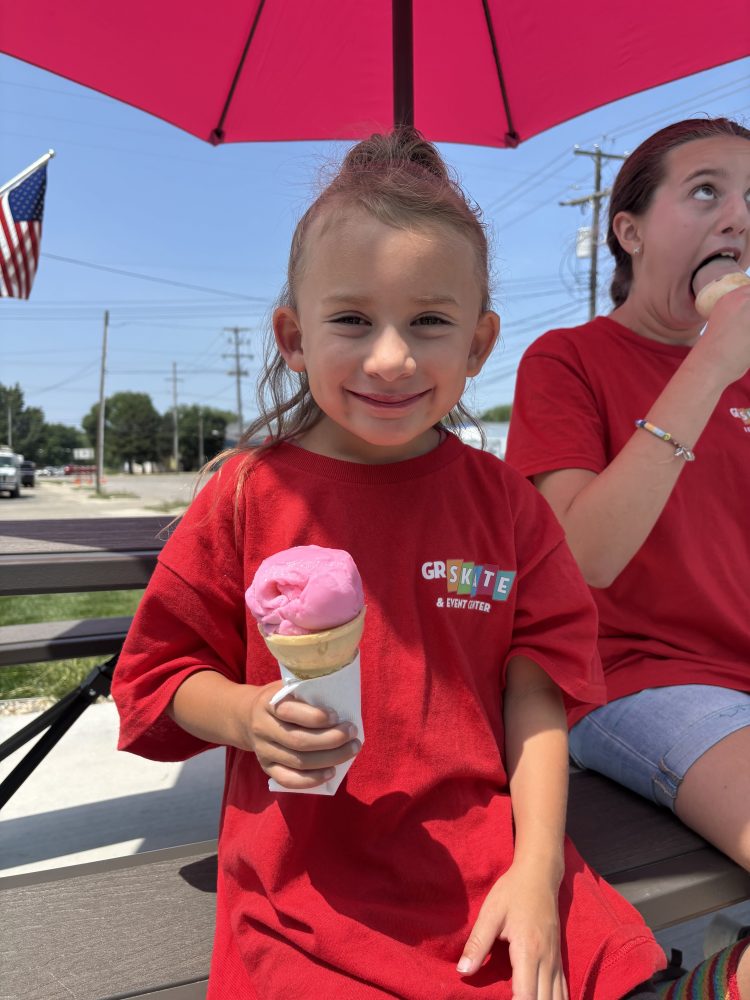 Two children in red shirts eating ice cream cones under a pink umbrella.