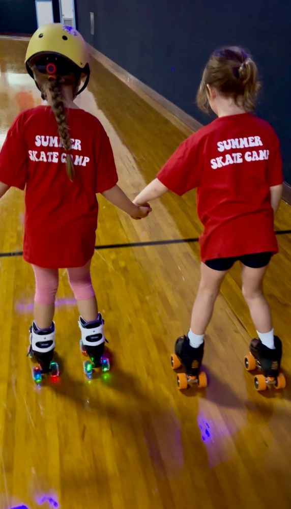Two kids in red shirts roller skating on a rink holding hands.