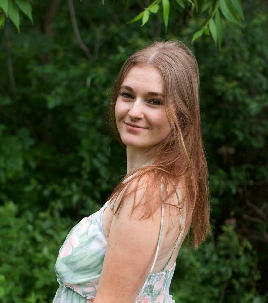 Woman with long hair in a floral dress standing outdoors, smiling.
