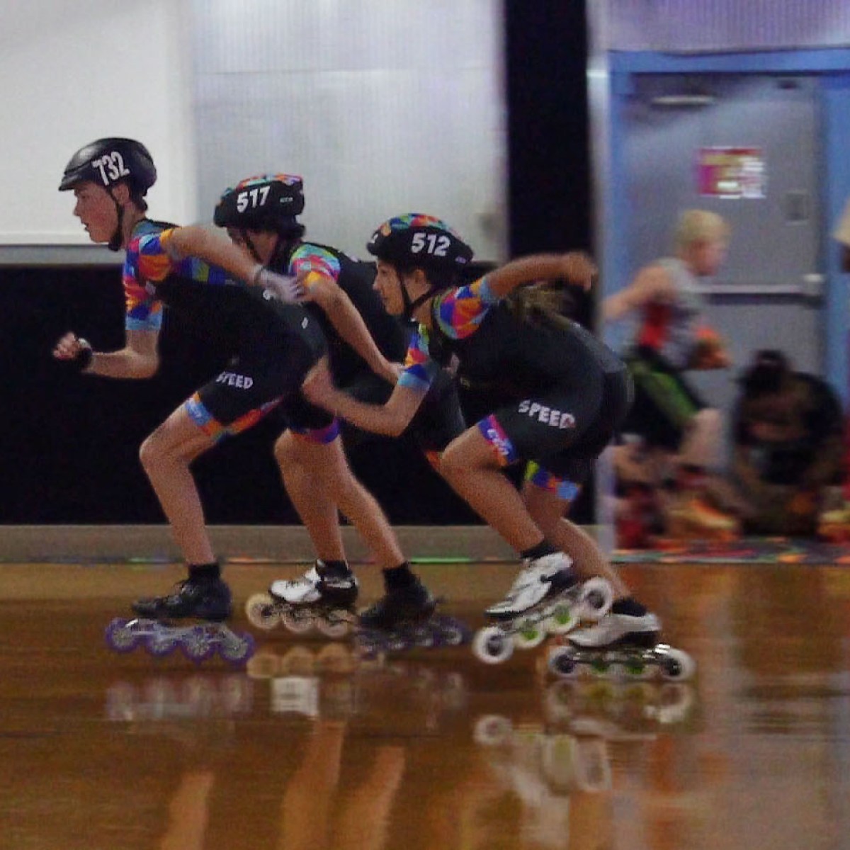 Three kids rollerblading indoors, wearing helmets and racing outfits.