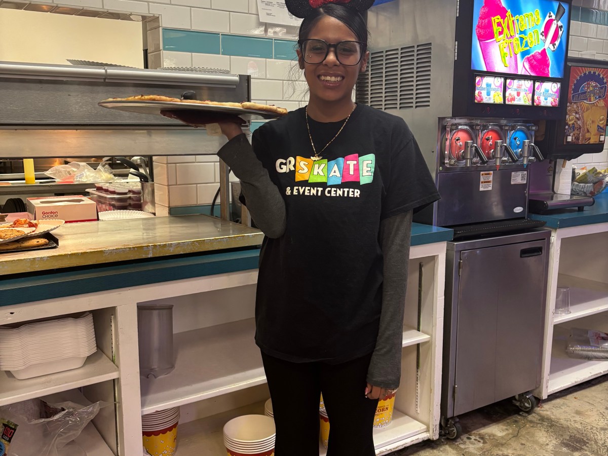 Person in mouse ears and apron holding trays in a food service area.