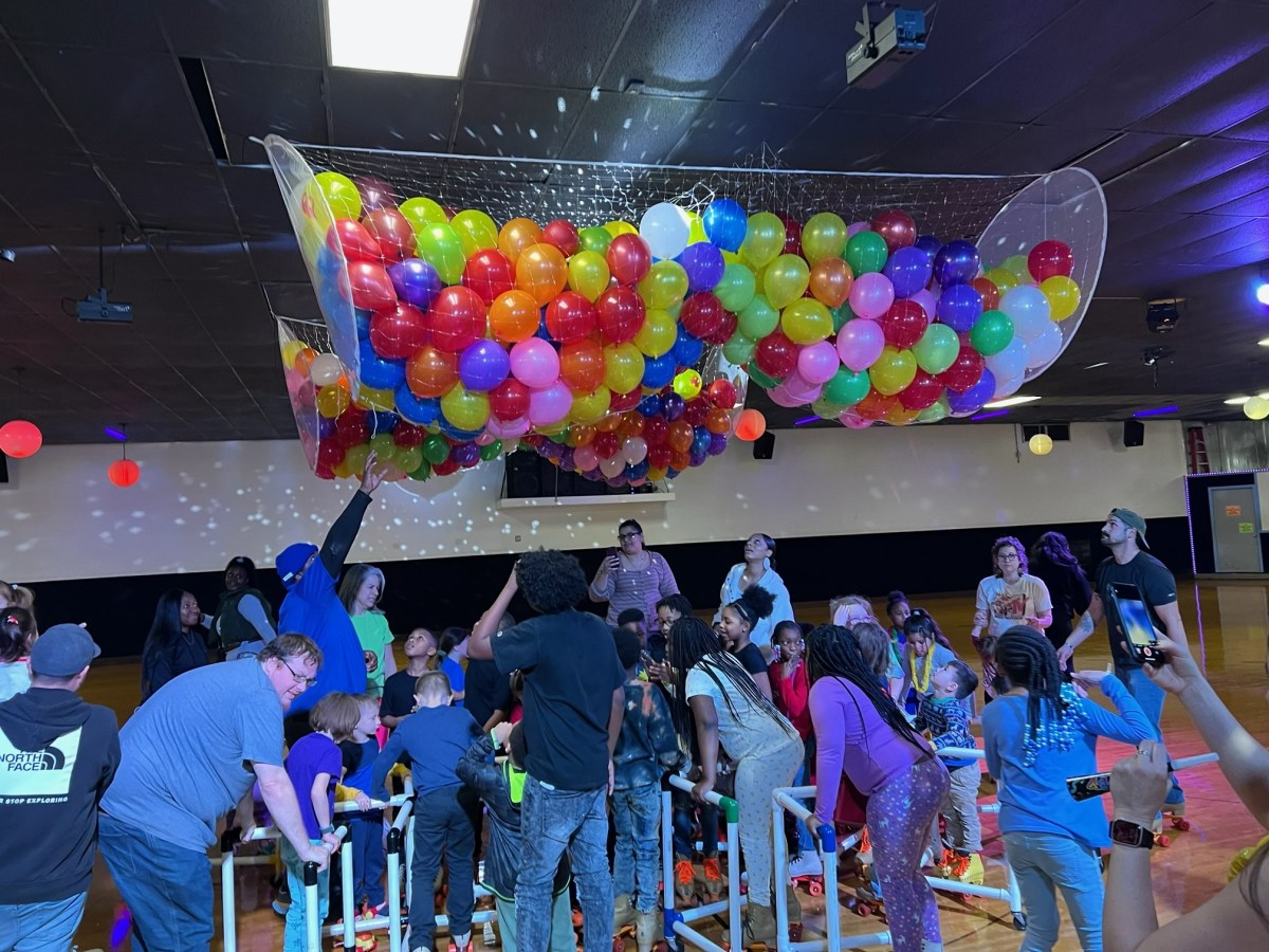 Group of people beneath a net filled with colorful balloons in a lit room.