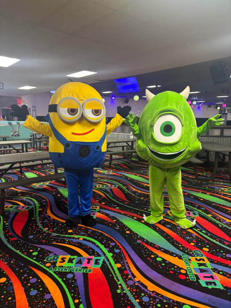 Two people in cartoon costumes: a yellow character and a green one-eyed character at a skating rink.