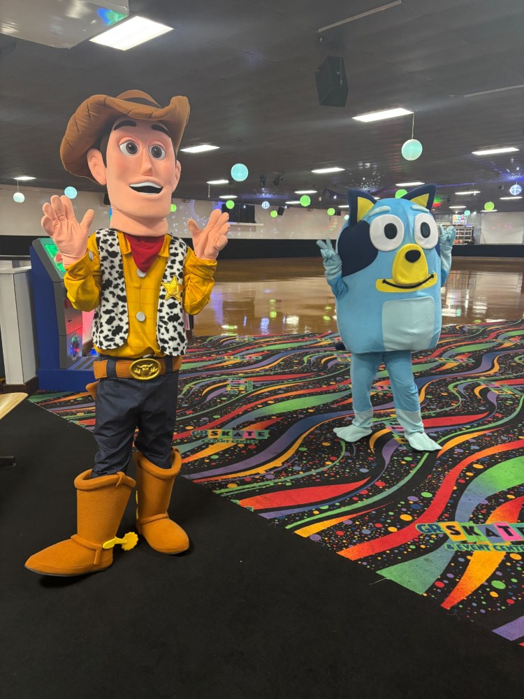 Two mascots, one cowboy and one blue dog, stand on a colorful rink floor.
